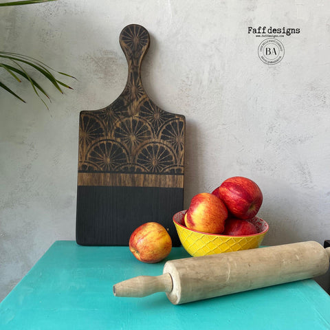 Belles and Whistles When Life Gives You Lemons - Stencil.  A wooden cutting board that is chalk painted black on the lower half and has the When Life Gives You Lemons stencil applied to the top and handle with black chalk paint.  A yellow and red bowl sits on the table holding red applies; an apple is sitting on the table in front of the cutting board; and a brown wooden rolling pin is sitting in front of the bowl.  All sits on a blue table, ion front of a gray wall.  Greenery sits at the left of the table.