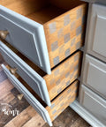 Belles and Whistles Whimsical Check - Stencil provides an extra decorative touch to any project!  Part of a gray chalk painted dresser with 3 drawers partially open.  The sides of all 3 drawers have the Whimsical Check stencil chalk painted on them in silver metallic paint.  Sitting on a multicolored brown wooden floor.