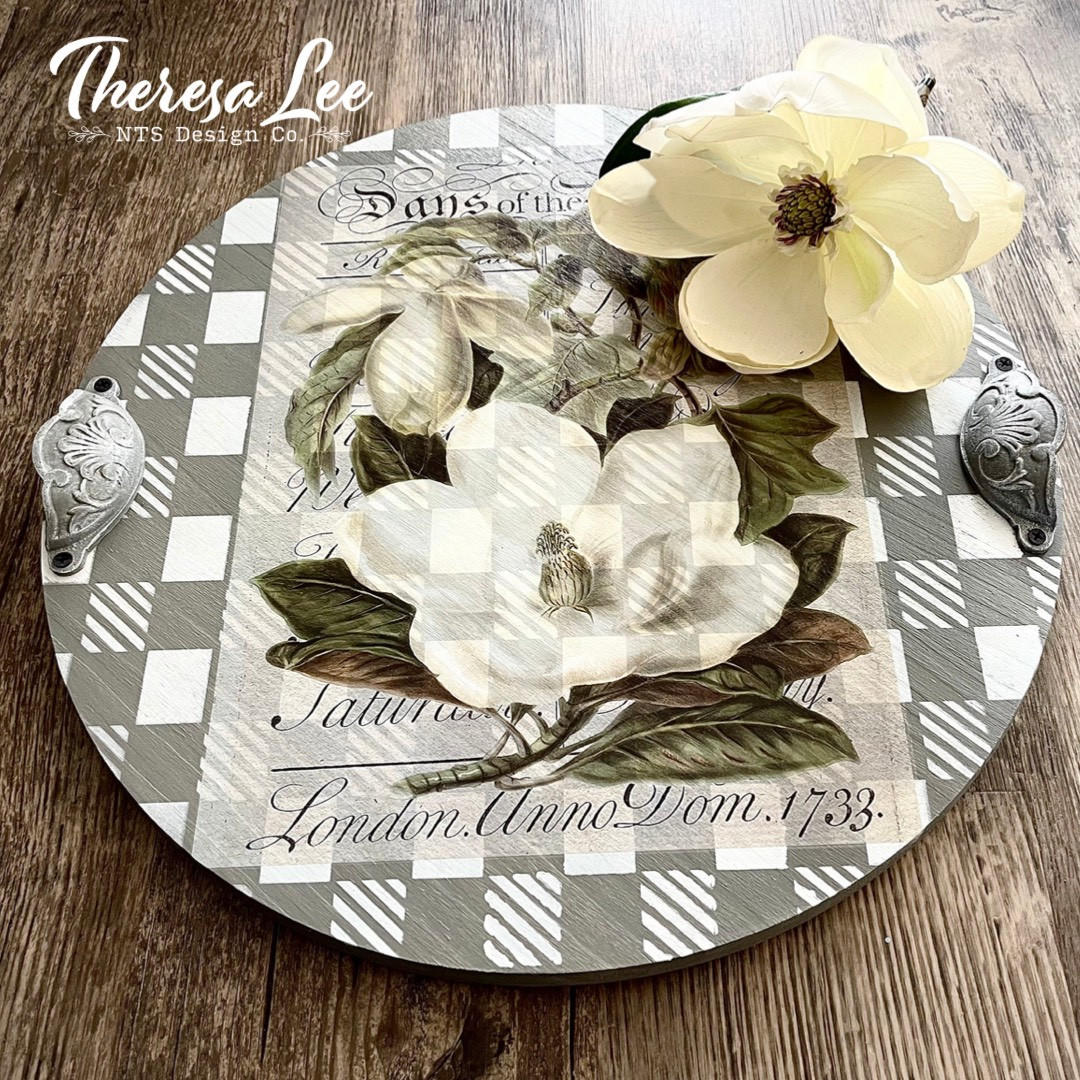 Belles and Whistles Whimsical Check - Stencil provides an extra decorative touch to any project!  A round serving tray that has the Whimsical Check stencil chalk painted on it in silver and white.  The Magnolia Garden transfer is applied to the top center of the tray, and the tray handles are silver.  The tray sits on a brown wooden table with a magnolia sitting on the upper right side of the tray.