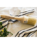 Belles and Whistles Best Dang Brush is a 2-inch blend that is excellent for painting, blending, and/or waxing. Image shows Best Dang Brush on top of white and blue striped cloth with greenery in the left corner.