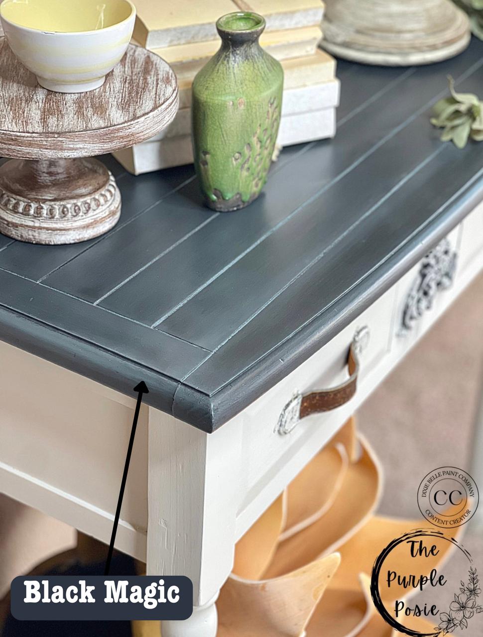 A rustic table with a stained black top labeled "Black Magic." The table has a white lower half with a brown leather handle, decorative vases, and stacked dishes on top. A logo in the corner reads "The Purple Posie.