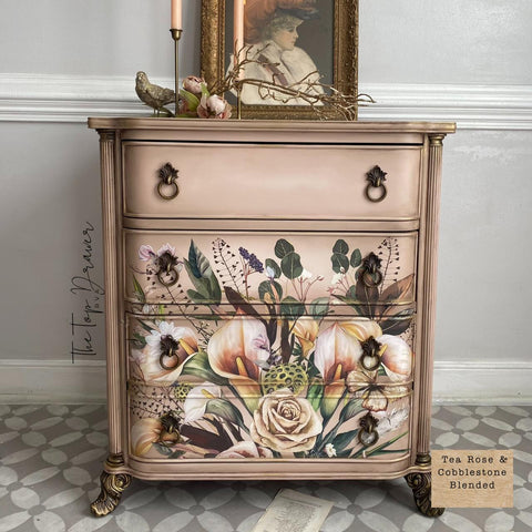 A painted dresser with a floral design features pale roses and greenery. A candle, framed art, and floral decor sit on top. The floor has a geometric pattern, and text reads "Tea Rose &amp; Cobblestone Blended" along with a small signature on the side.