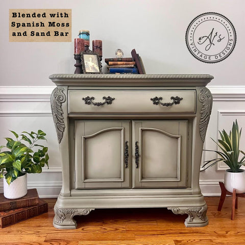 A vintage-style cabinet painted in shades of Spanish Moss and Sand Bar. It features ornate carvings, a drawer, and double doors. Decorated with books and small decor items on top, and houseplants on both sides against a white paneled wall.
