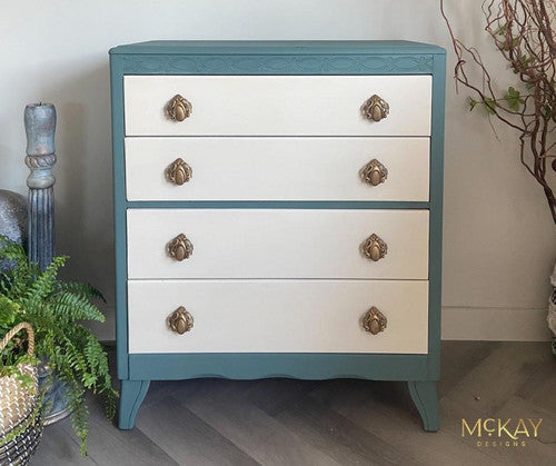 A vintage-style three-drawer chest with teal sides and white fronts featuring ornate brass knobs. The chest stands on curved legs, surrounded by a decorative vase and green potted plants. "McKay Designs" is in the bottom corner.