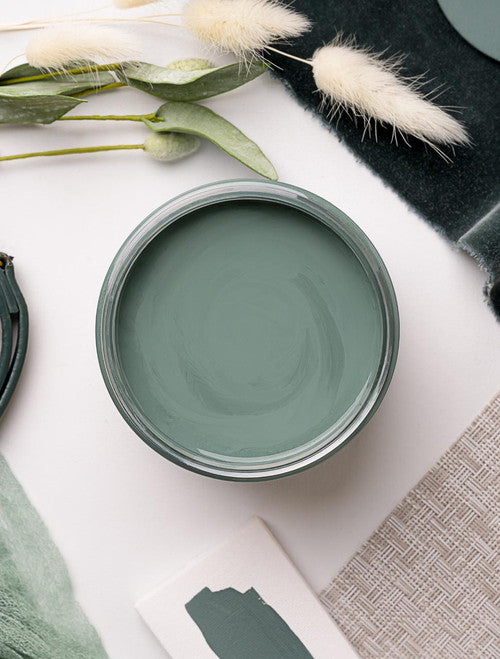 A flat lay of a green-themed arrangement featuring a jar of sage green paint, various shades of green fabric, a green purse, and sprigs of dried foliage on a white surface.