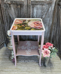 A vintage, shabby-chic style side table with floral motifs, painted in pastel pink and cream colors, elegantly standing against a painted rustic wooden backdrop with a bouquet of artificial flowers made using Blushing Roses A1 Rice Decoupage Paper.