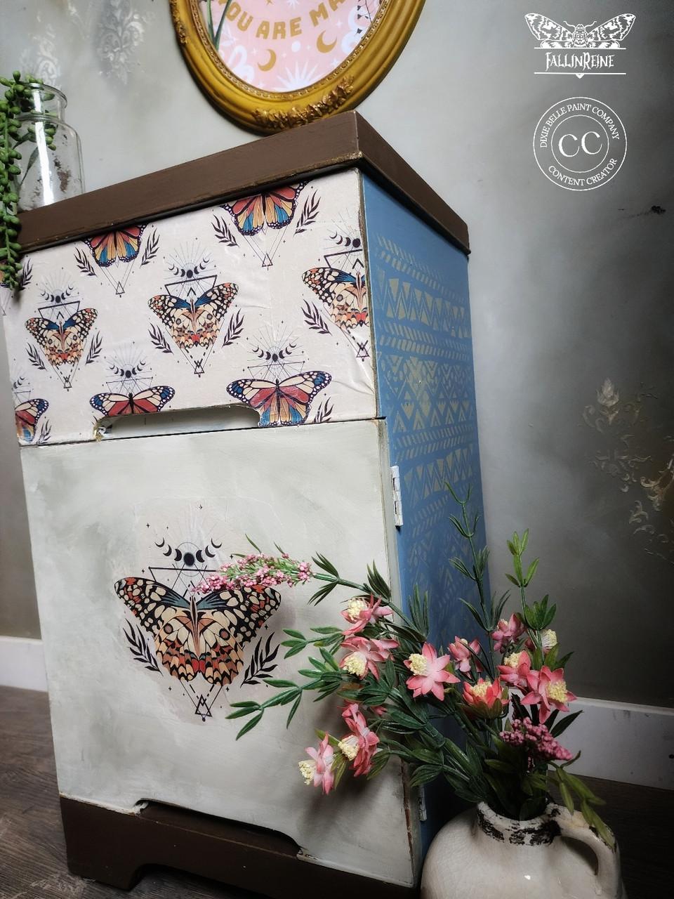 A two-drawer chest with butterfly designs on the front and floral patterns on the sides stands on a wooden floor. A vase with pink flowers is placed at its base. A round, gold-framed mirror hangs on the wall above.