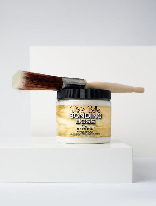 A jar of Dixie Belle Bonding Boss in clear, with a lid, sits on a white platform. A paintbrush with light bristles and a wooden handle rests on top of the jar against a plain white background.