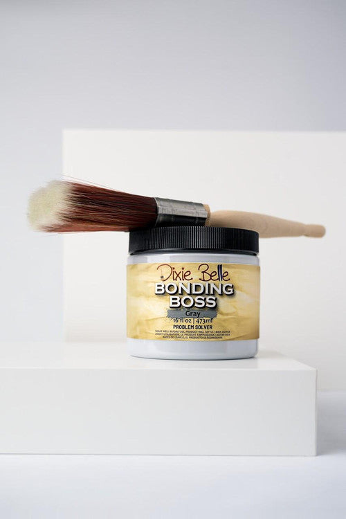 A paintbrush rests on top of a container of Dixie Belle Bonding Boss primer in gray, placed on a white geometric surface with a minimalist background.