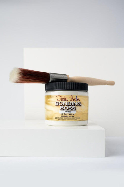 A jar of Dixie Belle Bonding Boss white primer sits on a white platform with a paintbrush resting horizontally on its black lid against a minimalist white background.