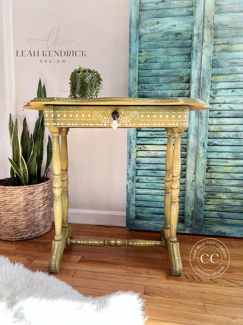 A vintage yellow entryway table with the Bone Inlay Stencil applied in decorative patterns with a crystal drop drawer pull, set against a pale blue rustic shutter backdrop, adorned with a small green plant on top, and complemented by indoor plants and a cozy white fur rug