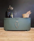 A vintage green chest with ornate metal detailing stands against a dark wall. On top, there's a black vase with dried flowers, a small blue bottle, and a wicker basket containing dried grass. The chest sits on a wooden floor.