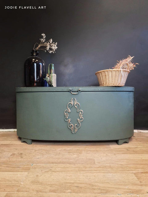 A vintage green chest with ornate metal detailing stands against a dark wall. On top, there's a black vase with dried flowers, a small blue bottle, and a wicker basket containing dried grass. The chest sits on a wooden floor.