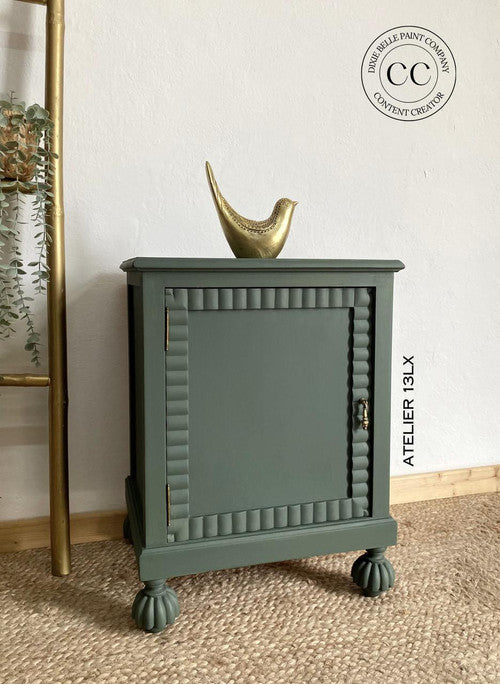 A small, green cabinet with decorative grooved edges and round feet sits on a woven rug. A golden bird figurine rests on top. The wall has a logo and plants nearby. A gold ladder is partially visible on the left.