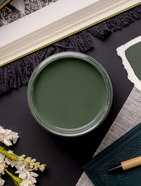 A top view of an open jar of dark sage green paint surrounded by fabric samples, a book, and white flowers on a dark surface. The arrangement suggests a color-coordinated design theme.