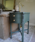 A green vintage side table with ornate details and two brass knobs stands beside a beige upholstered chair. A gold desk lamp is positioned on top. The background features a framed mirror and soft interior lighting.