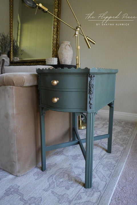 A green vintage side table with ornate details and two brass knobs stands beside a beige upholstered chair. A gold desk lamp is positioned on top. The background features a framed mirror and soft interior lighting.
