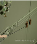 Close-up of a sage green cabinet with ornate detailing and bronze handles. A label pointing to the detail reads "Bronze Gilding Wax." Eucalyptus leaves hang on the left. Text at the bottom says "GRACIE'S HOUSE Painted Furniture.