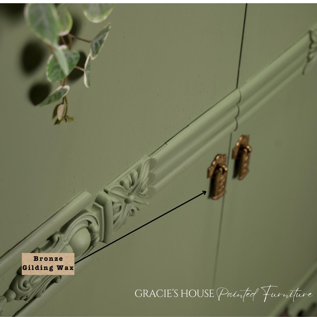 Close-up of a sage green cabinet with ornate detailing and bronze handles. A label pointing to the detail reads "Bronze Gilding Wax." Eucalyptus leaves hang on the left. Text at the bottom says "GRACIE'S HOUSE Painted Furniture.