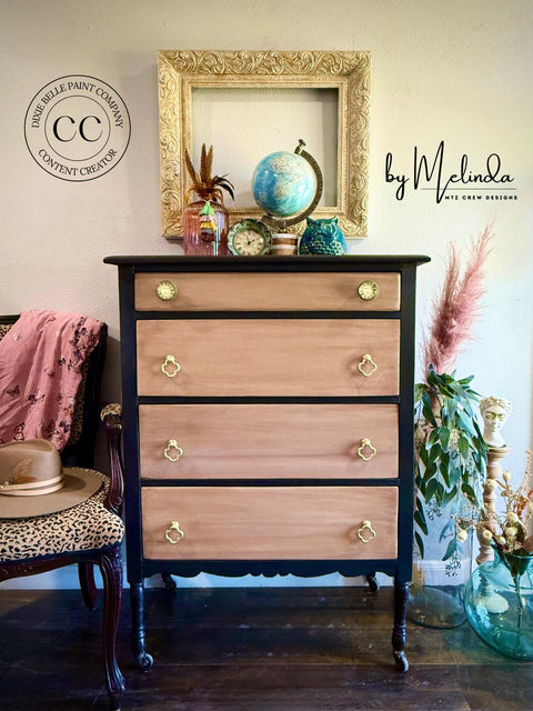 A vintage dresser with light wood drawers and dark frame is topped with books, globes, and a clock. An empty ornate frame hangs above. A plant, pink scarf, and hat decorate the scene. Logos on the wall read â€œDixie Belle Paint Company Content Creatorâ€ and â€œby Melinda MTZ Crew Designsâ€.