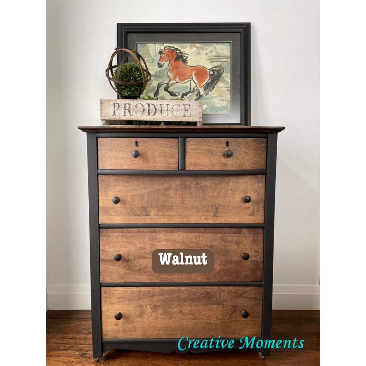 A wooden dresser with the word "Walnut" on one of the drawers. On top, there's a framed picture of a horse and a sign that reads "Produce." The floor is wooden, and the wall is plain white. "Creative Moments" is written in blue on the floor.