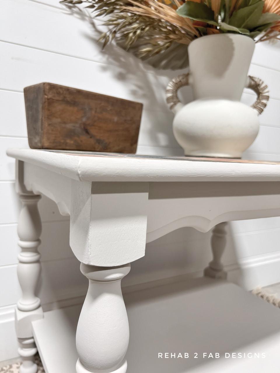 A white wooden side table with turned legs holds a rustic wooden box and a white vase with handles filled with dried foliage. The background features white shiplap walls. "REHAB 2 FAB DESIGNS" is written at the bottom.