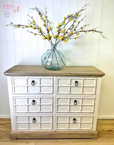 A white and wood dresser with six drawers stands against a white paneled wall. On top, there is a large clear glass vase holding long branches with yellow blossoms. The floor is light wood.