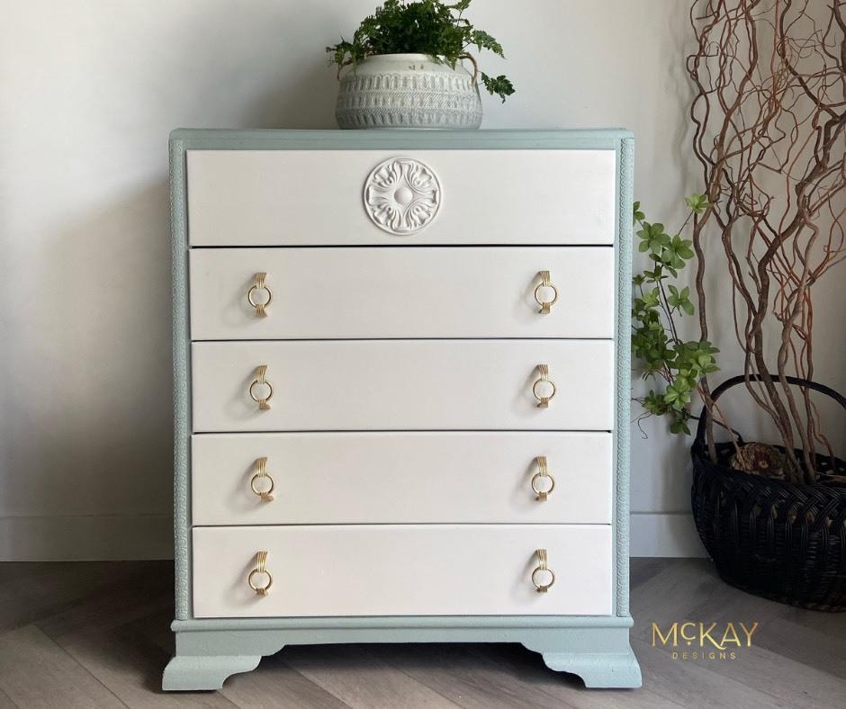 A small, five-drawer dresser with light blue edges and white drawers, featuring gold ring pulls. A white decorative medallion is on the top drawer. A potted plant sits on top, with another plant in a black basket nearby.