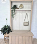 A light wood hall tree with a bench and upper shelf, decorated with a potted plant, trailing greenery, a golden decor piece, a hanging purse, and a small book against a white paneled wall and brick background.
