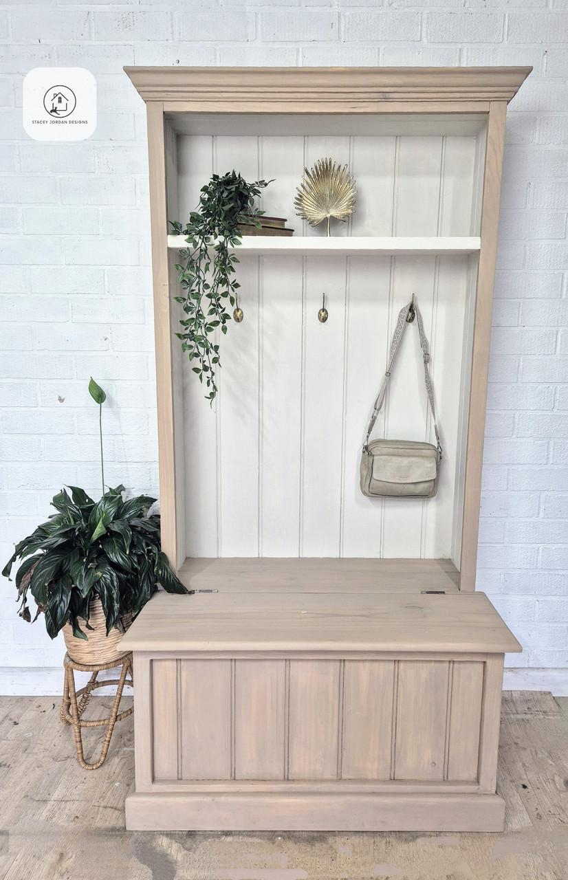 A light wood hall tree with a bench and upper shelf, decorated with a potted plant, trailing greenery, a golden decor piece, a hanging purse, and a small book against a white paneled wall and brick background.