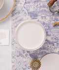 A jar of white paint on a lace tablecloth with a blue toile decoupage paper nearby, with part of a gold-rimmed saucer, brass handle, and decorative button visible nearby.