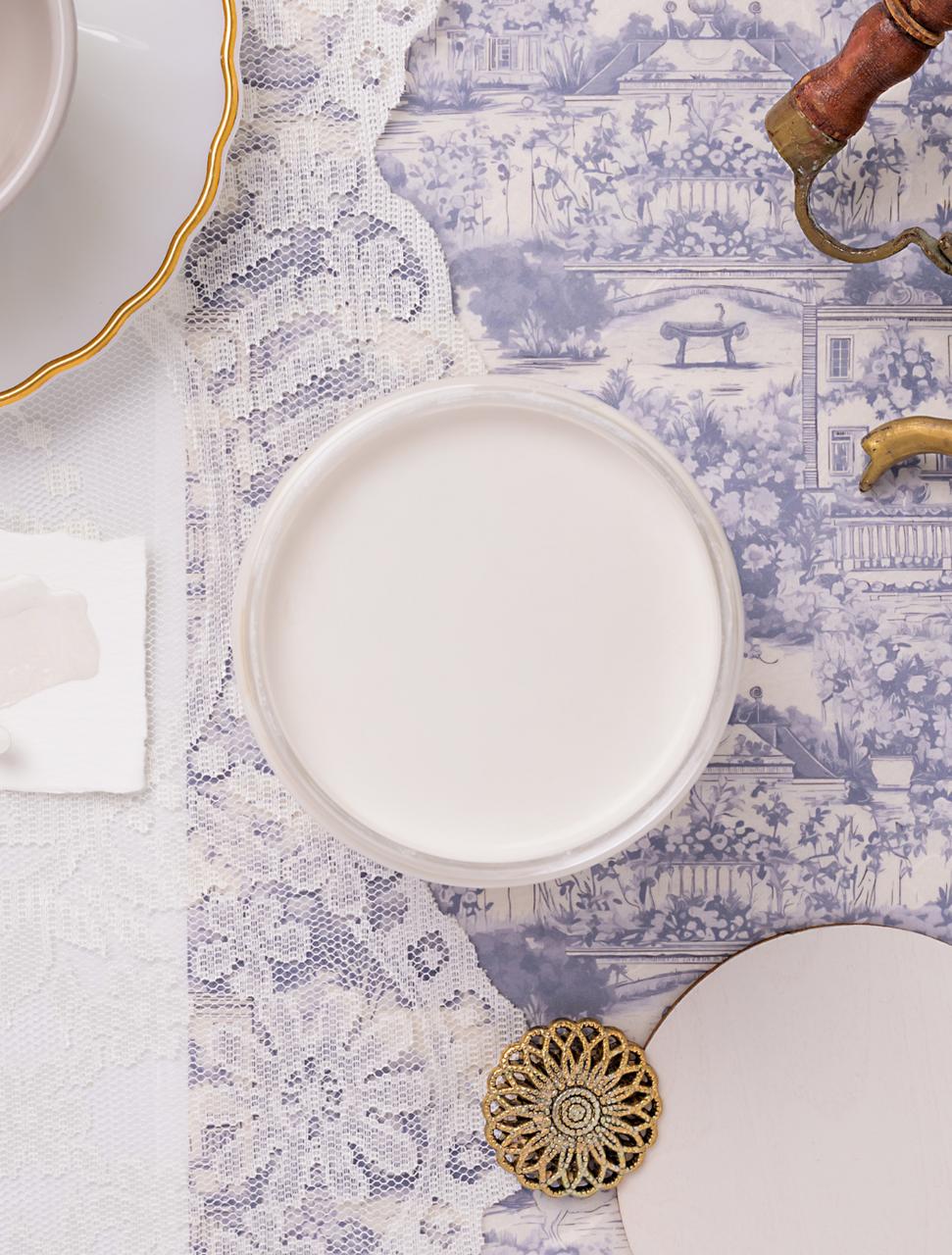 A jar of white paint on a lace tablecloth with a blue toile decoupage paper nearby, with part of a gold-rimmed saucer, brass handle, and decorative button visible nearby.
