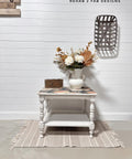 A white side table with floral detail holds a white vase of dried flowers and a wooden box, placed on a woven rug in front of a white shiplap wall with wall decor and the watermark "REHAB 2 FAB DESIGNS."