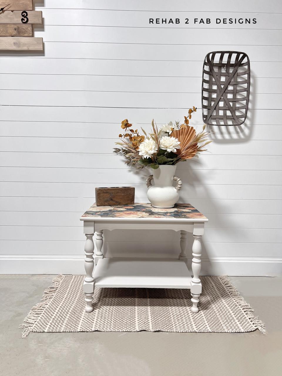 A white side table with floral detail holds a white vase of dried flowers and a wooden box, placed on a woven rug in front of a white shiplap wall with wall decor and the watermark "REHAB 2 FAB DESIGNS."