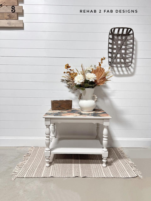 A white side table with floral detail holds a white vase of dried flowers and a wooden box, placed on a woven rug in front of a white shiplap wall with wall decor and the watermark "REHAB 2 FAB DESIGNS."