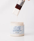 A hand holds a paintbrush dripping with white paint above an open container labeled "Silk All-In-One Mineral Paint, Chantilly" against a plain white background.