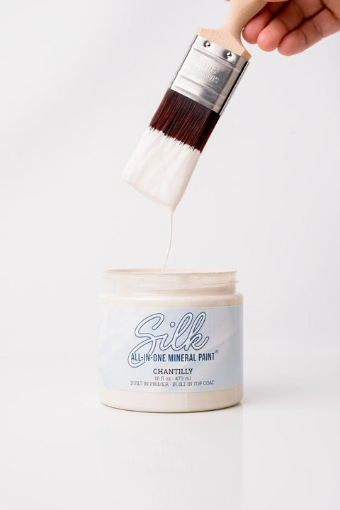 A hand holds a paintbrush dripping with white paint above an open container labeled "Silk All-In-One Mineral Paint, Chantilly" against a plain white background.
