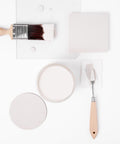 A flat lay of painting supplies on a white background, including a paintbrush with white paint, a palette knife, a square and round sample, and an open paint can with creamy white paint.