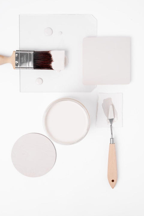 A flat lay of painting supplies on a white background, including a paintbrush with white paint, a palette knife, a square and round sample, and an open paint can with creamy white paint.