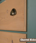 Close-up of a wooden cabinet drawer with a bronze handle, featuring a charred hickory finish. The side panel is painted teal. Text in the corner reads "Charred Hickory.
