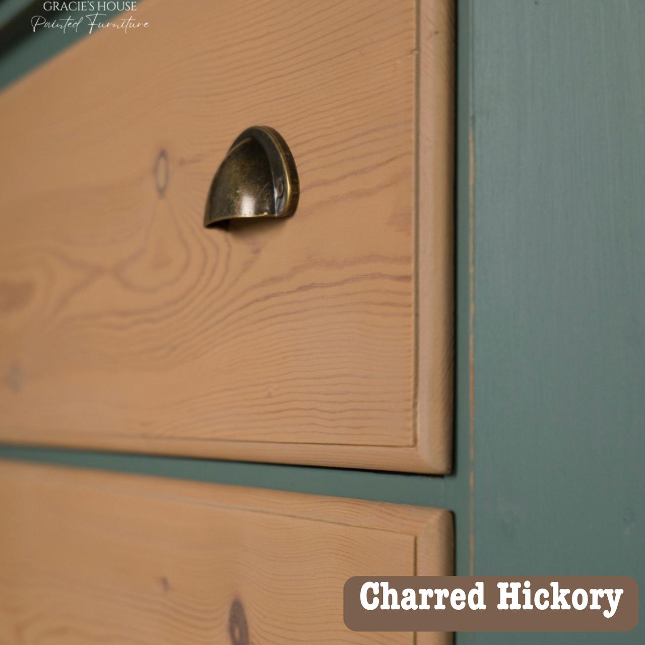 Close-up of a wooden cabinet drawer with a bronze handle, featuring a charred hickory finish. The side panel is painted teal. Text in the corner reads "Charred Hickory.