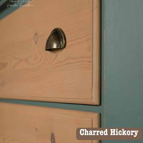 Close-up of a wooden cabinet drawer with a bronze handle, featuring a charred hickory finish. The side panel is painted teal. Text in the corner reads "Charred Hickory.