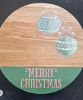 Wooden circular board with "Merry Christmas" from the Christmas Cheer stencil on the bottom applied in pink over green chalk paint. Two ornaments were applied with green, pink, purple, and white chalk paint from the Holiday Joy stencil.