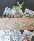 Long rectangular wooden board with the Christmas Cheer stencil's trees and snowflakes applied with white and green chalk paint. Antlers applied with brownish tan chalk paint and "Merry Christmas" applied with green chalk paint.