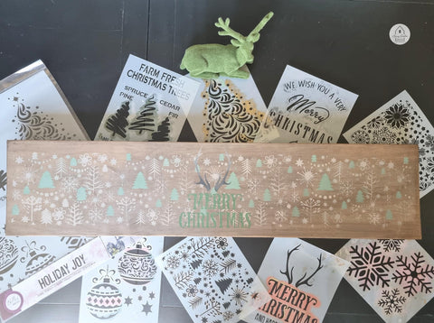 Long rectangular wooden board with the Christmas Cheer stencil's trees and snowflakes applied with white and green chalk paint. Antlers applied with brownish tan chalk paint and "Merry Christmas" applied with green chalk paint.