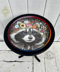 A round wooden table features a detailed painting of a raccoon wearing a colorful floral crown. The table's top has a black rim, and the legs are black as well. The background behind the table is white paneling, and logos are visible in the background.