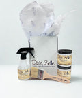 A gift box with tissue paper contains Dixie Belle products: a spray bottle, a paintbrush, and two small jars labeled "Clear Coat Satin" and "Chalk Mineral Paint Fluff", all against a plain white background.