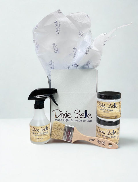 A gift box with tissue paper contains Dixie Belle products: a spray bottle, a paintbrush, and two small jars labeled "Clear Coat Satin" and "Chalk Mineral Paint Fluff", all against a plain white background.