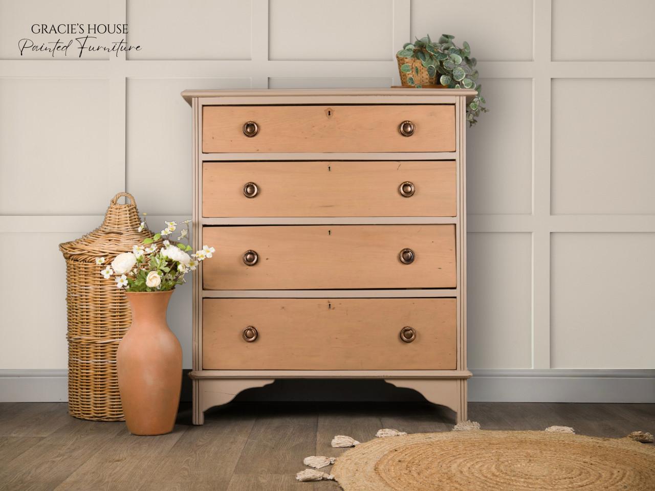A vintage-style wooden dresser with four drawers, placed against a paneled white wall. It is adorned with a small potted plant on top. Beside it, there's a terracotta vase with flowers and a woven basket on a wooden floor with a round rug.