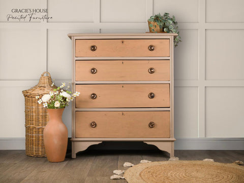 A vintage-style wooden dresser with four drawers, placed against a paneled white wall. It is adorned with a small potted plant on top. Beside it, there's a terracotta vase with flowers and a woven basket on a wooden floor with a round rug.